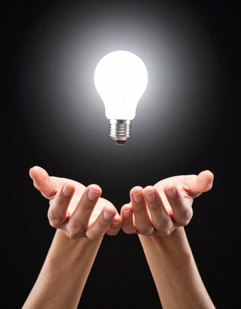 A pair of hands gently cradle a brightly glowing lightbulb against a dark background.の素材