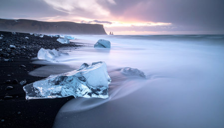 As dawn breaks over Iceland's dramatic coastline, waves gently wash over ancient glacial ice resting on the black volcanic sand of Diamond Beach.の素材