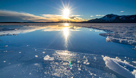 As the winter sun dips below the distant mountains, its golden light creates a brilliant starburst, casting a long, shimmering reflection across the surface of a partially frozen lake.の素材