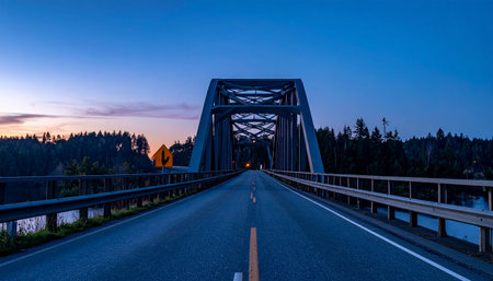 The road stretches forward, leading into the steel embrace of an arch bridge as twilight settles over the landscape.の素材