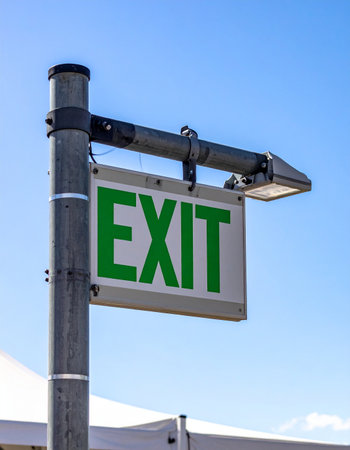 A clear green exit sign points the way forward against a bright, cloudless blue sky.の素材