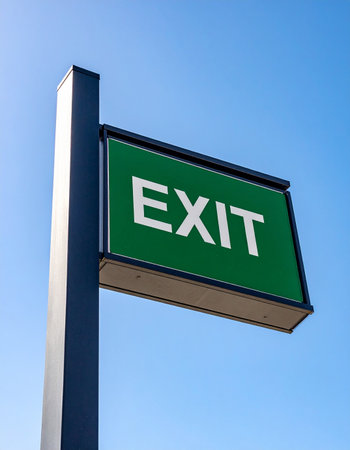 A bold green exit sign stands out against a vast, clear blue sky, offering clear direction and a sense of finality.の素材