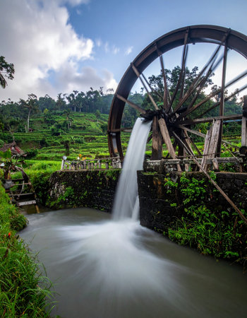 A traditional wooden water wheel turns endlessly, its motion captured in a smooth blur as it channels life-giving water to the vibrant green rice paddies below.の素材