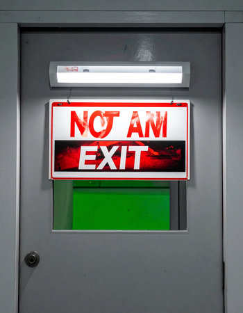 A brightly lit sign on a closed gray door offers a stark and confusing message: 'NOT AN EXIT'.の素材
