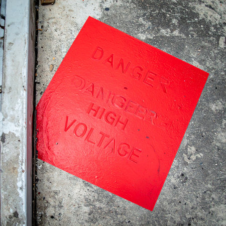 A weathered red sign lies on a gritty concrete floor, its embossed letters serving as a stark and urgent warning of the unseen electrical hazard nearby.の素材