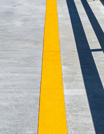 A bold yellow line cuts across a gray concrete surface, creating a strong visual path. Bright sunlight casts long, dark shadows, adding a dramatic, graphic quality to the scene.の素材