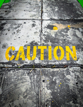 A stark yellow 'CAUTION' stencil is painted across a worn and dirty industrial floor, serving as a clear warning.の素材