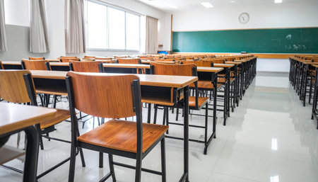 Rows of empty wooden desks and chairs sit in a quiet, sunlit classroom, patiently awaiting the arrival of students.の素材