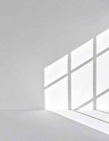 Morning light streams through a window, casting a grid of shadows across the clean white walls and floor of an empty room.の素材