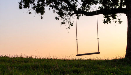 An empty swing hangs from the branch of a large tree, silhouetted against the warm, gentle glow of a setting sun.の素材