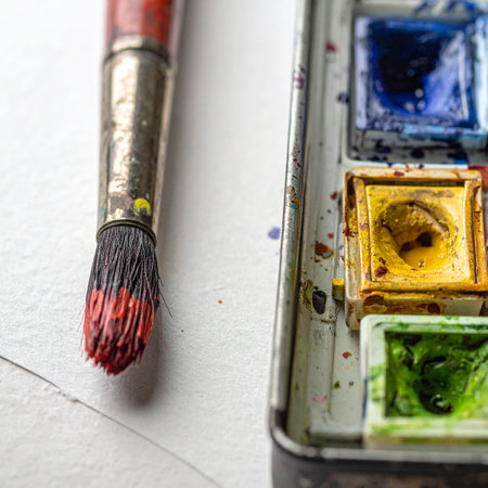 A well-used paintbrush, its bristles stained with vibrant red, rests beside a watercolor palette, hinting at a creative session in progress.の素材