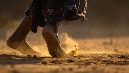 A low-angle, close-up shot captures the dynamic energy of a child's bare feet running across a dusty path.の素材