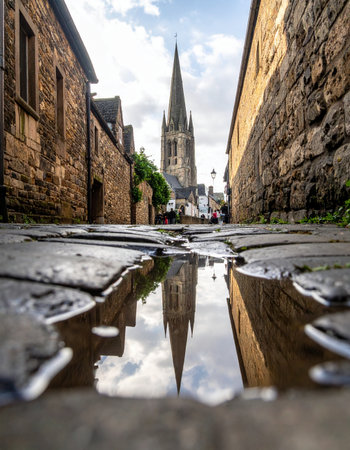 A dramatic low-angle view captures the magnificent spire of a historic European church, its perfect reflection mirrored in a rain puddle on an ancient cobblestone street.の素材