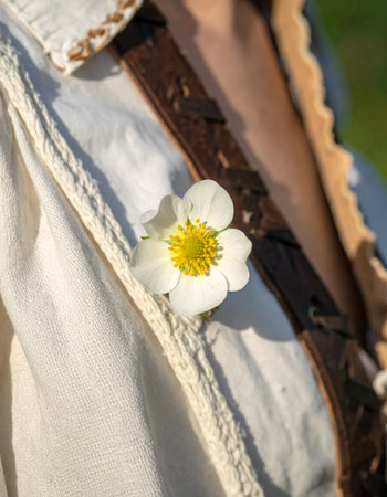 A single white wildflower, freshly picked from a sun-drenched meadow, is tucked into a rustic strap against a soft linen dress.の素材