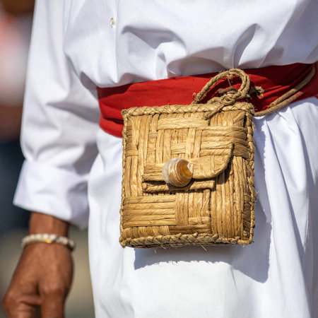 A close-up detail shot captures a handcrafted woven bag, made from natural fibers, attached to a vibrant red sash.の素材