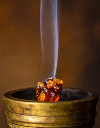 A single piece of glowing amber resin smolders in an antique brass censer, releasing a fragrant plume of white smoke.の素材