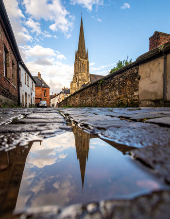 A low-angle view captures the magnificent spire of St James' Church in Louth, perfectly reflected in a rain puddle on a historic cobblestone lane.の素材
