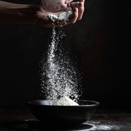 In the quiet focus of a dimly lit kitchen, a chef's hands carefully sift fine white flour, a cloud of powder cascading into a dark bowl.の素材