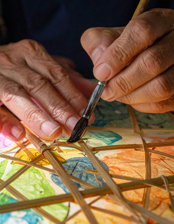 In a sunlit workshop, the weathered hands of a master artisan delicately apply vibrant watercolors to a traditional paper lantern.の素材