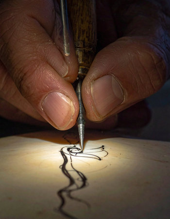 In a warmly lit, focused moment, an artist's steady hand guides a traditional dip pen across paper.の素材