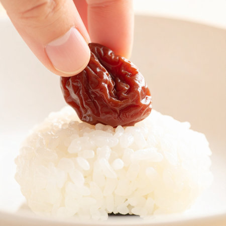 A close-up shot captures the careful placement of a traditional Japanese umeboshi, or pickled plum, onto a bed of fresh white rice.の素材