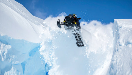 An expert rider launches their high-performance snowmobile off a snowy ledge, catching massive air against a brilliant blue sky.の素材