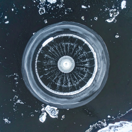 An aerial perspective reveals a perfectly formed ice circle spinning slowly in the dark, frozen waters of a remote lake.の素材