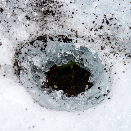 A close-up view of a circular hole melting through a layer of dirty snow and ice, revealing the dark, mysterious water below.の素材