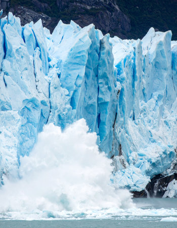 A colossal wall of ancient blue ice fractures with a thunderous roar. A massive iceberg breaks free and crashes into the frigid water below, sending up a powerful splash.の素材