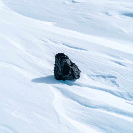 A single, dark rock rests in stark contrast against a pristine, windswept field of white snow.の素材