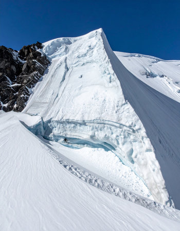 A breathtaking view up a steep, snow-covered mountain face towards a sharp summit under a clear blue sky.の素材
