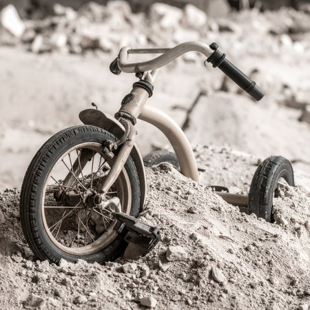 A poignant black and white photograph of a forgotten child's tricycle, broken and half-buried in a mound of sand and rubble.の素材