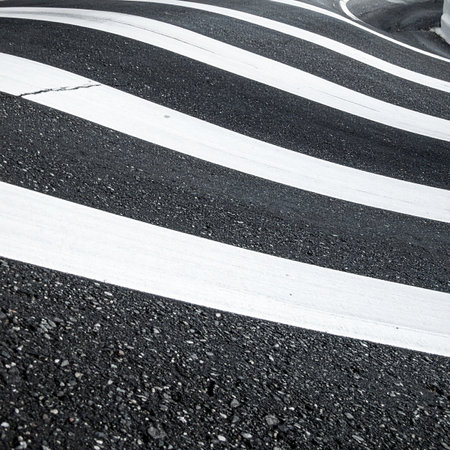 From a low angle, the bold white lines of a zebra crossing create a dynamic, wave-like pattern across the textured black asphalt.の素材