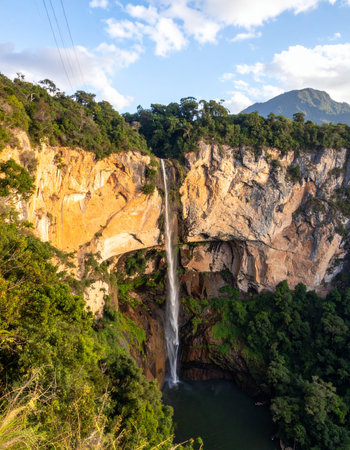 A breathtaking aerial view captures the immense scale of a powerful waterfall as it plunges hundreds of feet down a sheer, sun-kissed cliff face into a hidden pool below.の素材