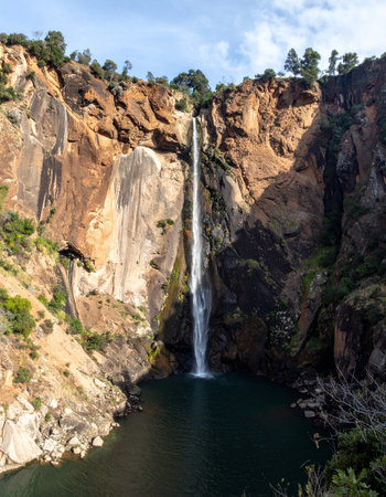A powerful stream of water carves its path down a sheer, sun-kissed rock face, plunging into the tranquil, dark pool below.の素材