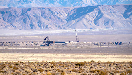 In the vast, sun-scorched expanse of a remote desert, an oil pumpjack tirelessly works, a solitary silhouette against a backdrop of hazy mountains.の素材