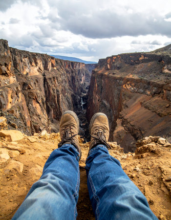 A moment of rest and reflection after a long hike. From a first-person perspective, a hiker's boots dangle over the edge of a vast, rocky canyon, taking in the breathtaking and dramatic landscape.の素材