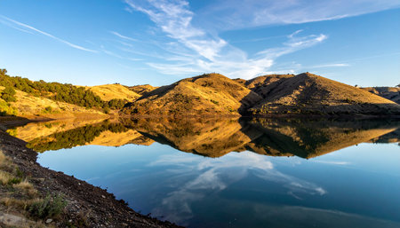 A breathtaking panoramic view of rolling golden hills perfectly mirrored in the still, glassy surface of a tranquil lake.の素材