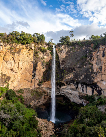 A breathtaking view of a slender waterfall as it makes its dramatic descent from a sun-drenched cliff.の素材