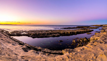 The sky ignites in a gradient of purple, pink, and gold as the sun sets over a rugged, rocky coastline.の素材