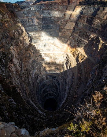 Sunlight catches the upper terraces of a colossal open-pit mine, highlighting the immense scale of human engineering.の素材