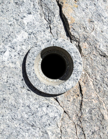 A perfectly circular hole drilled with industrial precision into a raw, cracked granite surface. A crescent shadow plays across the stone, creating a mysterious and abstract symbol of contrast between nature and technology, evoking concepts of depth and exploration.の素材