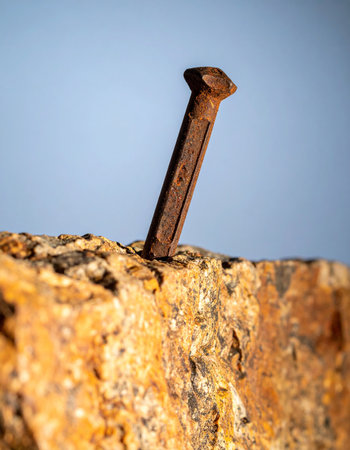 An old, rusty nail stands firm, embedded in a weathered piece of stone against a clear blue sky. This macro shot captures the texture of corrosion and rock, symbolizing strength, resilience, and the enduring passage of time. A powerful metaphor for perseverance and history.の素材