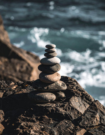 A carefully constructed stack of smooth stones stands in perfect balance on a rugged cliff, a symbol of stability and mindfulness against the backdrop of restless ocean waves. This serene scene captures a moment of peace and harmony found in natures raw beauty.の素材