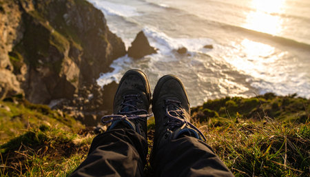 From a first-person perspective, a hiker takes a well-deserved rest on a grassy cliff, their boots pointing towards the magnificent view. Below, waves crash against the rugged coastline as the setting sun casts a warm, golden glow over the water, creating a moment of pure serenity and connection with nature.の素材