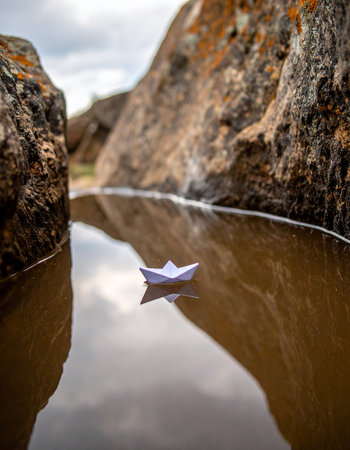 A single white paper boat floats serenely on the still, reflective surface of a hidden pool, navigating a narrow passage between ancient, weathered rocks. This quiet scene evokes feelings of peace, solitude, and the beginning of a gentle journey into the unknown.の素材