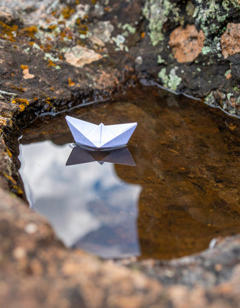 A tiny, handmade paper boat embarks on its first great voyage across a small puddle nestled among rugged rocks. Its reflection hints at the vast sky above, symbolizing the start of a grand adventure, fueled by imagination, hope, and the simple joys of childhood dreams.の素材