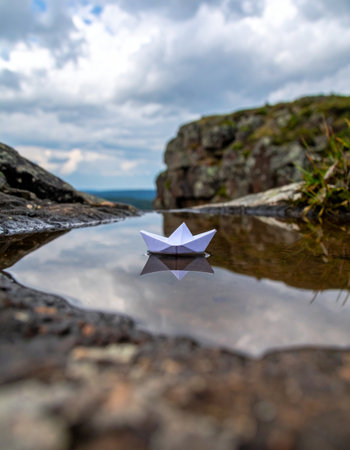 A single white paper boat embarks on a journey across a still, reflective puddle nestled among ancient rocks. The dramatic cloudy sky is mirrored in the water, suggesting a grand adventure awaits this small, fragile vessel. A symbol of dreams, hope, and the start of a new voyage.の素材