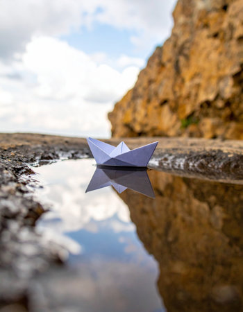 A small, handmade paper boat embarks on a grand voyage across a reflective puddle. With a backdrop of rugged cliffs and a vast sky, this simple toy represents big dreams, the start of an adventure, and the power of imagination in the face of natures scale.の素材