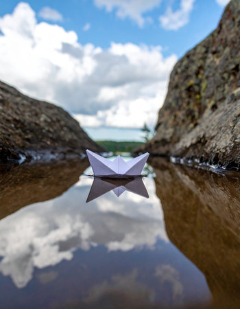 A tiny origami boat embarks on a grand adventure, navigating the calm, reflective waters between two rocky shores. It symbolizes a journey of hope, imagination, and the start of a new voyage into the unknown, with a vast sky of possibilities reflected below.の素材